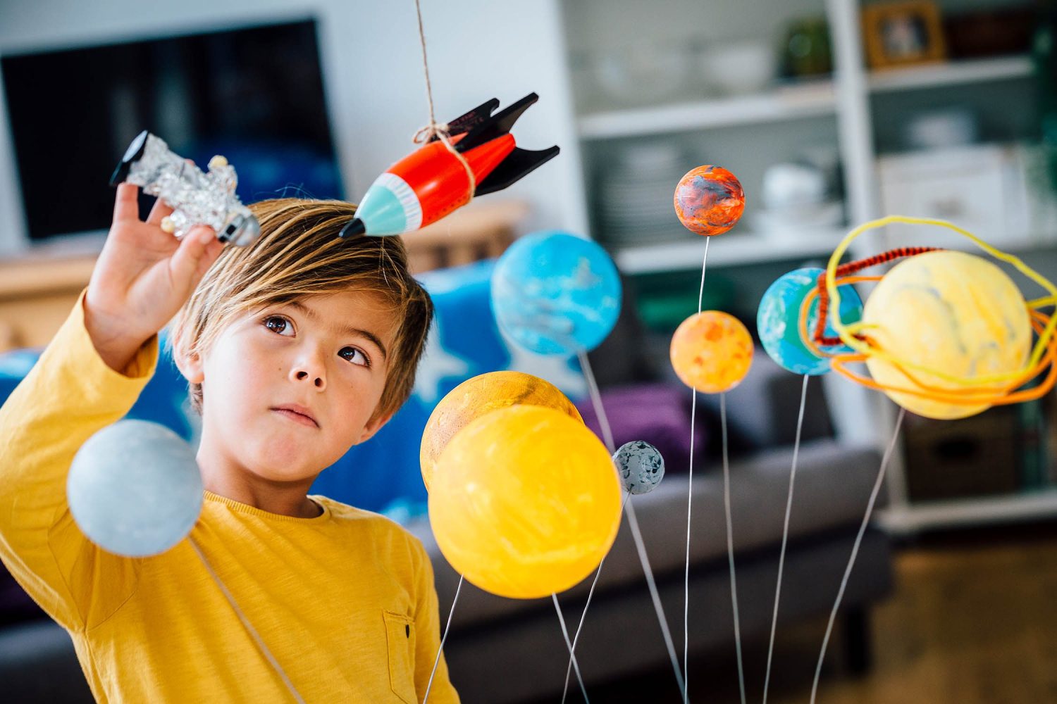 Little boy playing with his homemade planetarium as he holds an astronaut. A rocket hangs above. Arms raised as he plays.